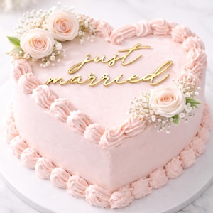 May include: A heart-shaped pink cake decorated with buttercream frosting and the words "just married" in gold script. The cake is adorned with pink roses and small white flowers, perfect for a wedding celebration.