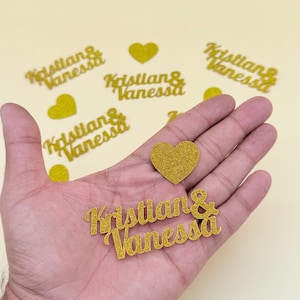 Può includere: Ritagli dorati glitterati dei nomi "Kristian & Vanessa" e forme di cuore. I nomi sono in corsivo. Diversi ritagli sono sparsi su una superficie giallo chiaro. Un cuore e un ritaglio di nome sono in una mano.