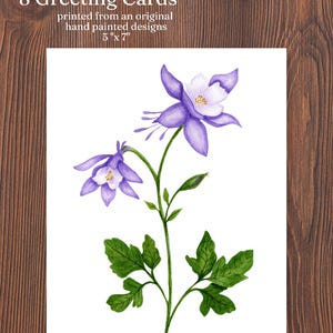 May include: A set of 8 greeting cards featuring a watercolor illustration of a purple and white columbine flower with green leaves and stems. The text on the card reads 8 Greeting Cards printed from an original hand painted designs 5" x 7".