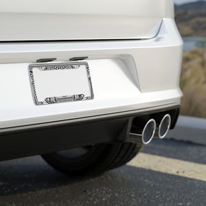 May include: A white car with a black and white patterned license plate frame. The car has dual exhaust pipes.