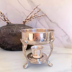 May include: A silver-plated metal warming stand featuring a circular bowl supported by three ornate, curved legs with claw-style feet. A small, matching silver-colored cup sits on a circular base plate positioned between the legs of the stand.