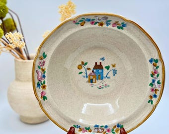 Può includere: Una ciotola da portata in ceramica beige con bordo smerlato e bordo marrone. La ciotola presenta un'illustrazione centrale di una casa blu e gialla con un bordo floreale e una farfalla. Il bordo è decorato con fiori e frutta colorati.