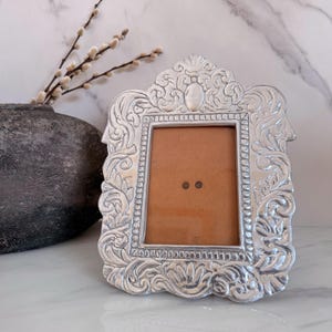 May include: A silver-toned picture frame with an ornate, embossed scrollwork border featuring intricate leaf and floral patterns. The rectangular frame has a central opening with a tan backing and two small, dark circular fasteners visible.