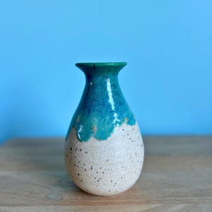 May include: A handcrafted ceramic vase with a speckled, off-white base and a turquoise and teal glazed top. The vase has a rounded body and a narrow neck. The vase is displayed on a wooden surface against a blue background.