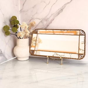 May include: A rectangular mirror with a gold-colored frame and a decorative stand. The mirror has a light-colored surface with a gold-colored pattern. A white vase with green and white flowers is to the left of the mirror.