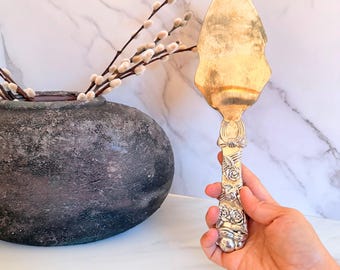 May include: A silver-toned metal cake server featuring a pointed, flat blade and an ornate handle decorated with embossed floral rose patterns. The handle transitions into a detailed neck design, providing a decorative aesthetic for serving.