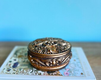 May include: A detailed, antique-style, bronze-colored trinket box with ornate floral and leaf designs. The box is round with a decorative lid. It rests on a book titled "Wind Flowers".