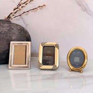 May include: Three photo frames: a rectangular silver frame with a beaded border, a rectangular gold frame with corner shell-like accents, and an oval gold frame with small ball feet. The gold frames contain inserts with text including 3x5 inches.