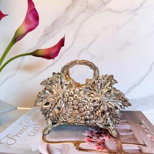 May include: A silver-toned metal napkin holder featuring a detailed embossed design of grape clusters and vine leaves. The holder has a central circular handle at the top and stands on small feet, showcasing a metallic finish with reflective highlights.