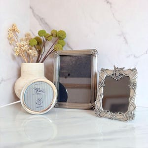 May include: Three silver-plated picture frames of varying shapes and sizes. A round frame, a rectangular frame, and a decorative frame with floral and angel accents. A vase with dried flowers is in the background.