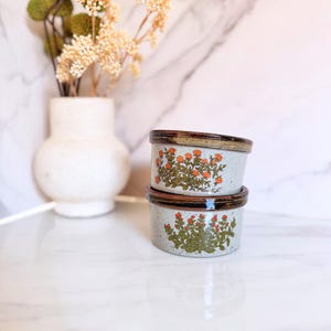 May include: Two stacked ceramic ramekins with a speckled light gray finish and dark brown rims. The top ramekin features an orange floral pattern, while the bottom ramekin displays a green and red botanical design with small berries.