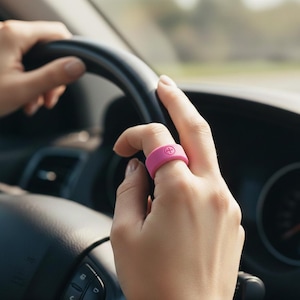 Puede incluir: Un anillo de silicona rosa brillante con un símbolo de suma se lleva en un dedo. La mano sostiene un volante negro. El fondo está borroso, lo que sugiere el interior de un coche.