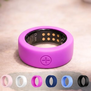 Puede incluir: Un anillo inteligente morado con un símbolo de suma. El anillo se muestra con otras opciones de color: rosa, blanco, magenta, azul marino, azul claro y negro. El interior del anillo es negro con pequeños puntos dorados. Un accesorio tecnológico portátil.