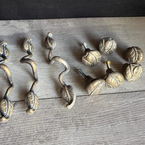 May include: A collection of four elongated, winding metal drawer pulls and four round metal knobs, all featuring a textured, antique brass finish with embossed leaf motifs. The hardware pieces are arranged in two groups on a light gray surface.