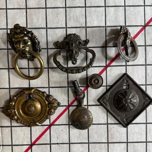 May include: A collection of six metal furniture hardware pieces including a lion head ring pull, an ornate oval pull, a small circular ring pull, a decorative round escutcheon, a pendant pull with a circular drop, and a diamond-shaped backplate.