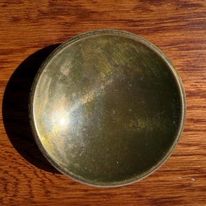 May include: A circular brass dish with a polished, reflective surface showing subtle signs of wear and fine scratches. The object features a slightly concave center and a smooth, rounded rim, exhibiting a warm, metallic golden-brown patina.