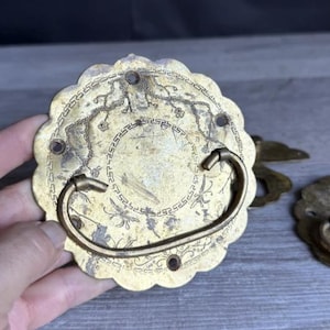 May include: A round brass drawer pull with a scalloped edge and a swinging handle. The surface features an engraved floral and geometric border pattern with a central circular motif. There are four small holes for mounting screws around the perimeter.