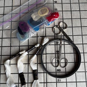 May include: A collection of embroidery supplies including white thread, scissors, a hoop, and a variety of colorful threads and buttons. The items are arranged on a grid-patterned surface, suggesting a crafting or sewing project.