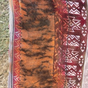 May include: Textile with a vibrant pattern, featuring orange, brown, and red hues. The design includes a tiger stripe-like pattern, geometric shapes, and a decorative border. The fabric is held by a clothespin.