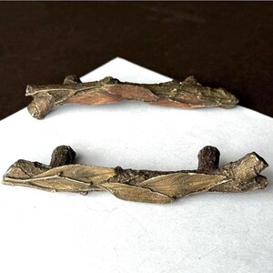 May include: Two cabinet pulls shaped like textured tree branches with attached leaves. The pulls feature a metallic finish with dark brown and muted gold tones, showcasing a realistic bark texture and elongated leaf details along the horizontal bar.