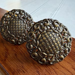 May include: Two round metal drawer knobs with an antique brass finish. Each knob features a central circular area with a raised, textured checkerboard pattern, surrounded by an intricate, openwork border of swirling, scroll-like metal filigree.