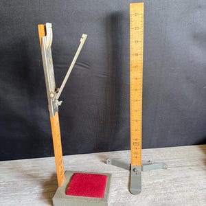 May include: Two vintage wooden and metal measuring tools. One tool has a red felt square and a hinged arm. The other is a wooden ruler with measurements up to 21 inches. Both tools are gray and orange.