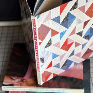 May include: A stack of books with the title "THE FAMILY CREATIVE WORKSHOP" visible on the spines. The top book is open, displaying pages and a cover with a geometric pattern in red, blue, and white. Other books have colorful covers.