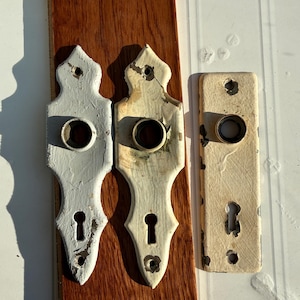 May include: Three metal door plates with chipped white paint. Two plates feature a pointed, decorative shape with a central circular opening and a keyhole. The third plate is rectangular with rounded corners, a central circular opening, and a keyhole.
