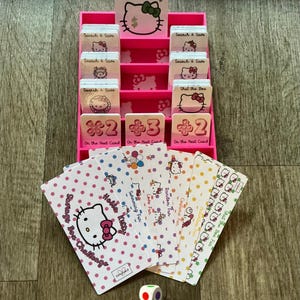 May include: A pink Hello Kitty-themed game set with a tiered card holder, cards, and a die. The card holder has multiple slots with cards that say "Search & Save" and "Shut the Box". The cards feature Hello Kitty designs.