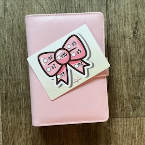 May include: A light pink planner with a matching bow-shaped sticker. The sticker has a pink bow design with various dollar amounts, including $2, $3, $5, $7, $8, and $10. The planner is closed and has a strap closure.