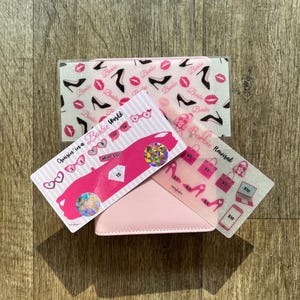 May include: A pink wallet with three clear overlays featuring Barbie-themed designs. The overlays display images of high heels, lipstick, and a pink car. Text includes "Cruisin' in a Barbie World" and "Barbie Revised".