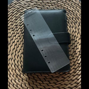 May include: Black leather planner with a clear plastic page marker. The planner has a snap closure and is set on a woven, circular placemat. The page marker has the word "HOLIDAYS" printed on it.