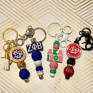 Op de afbeelding: Een verzameling sleutelhangers met verschillende ontwerpen. Sleutelhangers zijn onder meer een goudkleurige met een witte kwast, een blauw-witte met de letters ZΦB en een roze-groene met de letters AKA 1908. Andere sleutelhangers hebben Griekse letters.