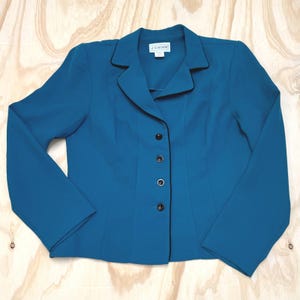 Puede incluir: Un blazer de color azul cerceta con solapa de muesca, mangas largas y cierre frontal de cuatro botones negros. La prenda presenta un fino ribete negro a lo largo de la solapa y el borde delantero, con una etiqueta interior que dice J.G. HOOK.