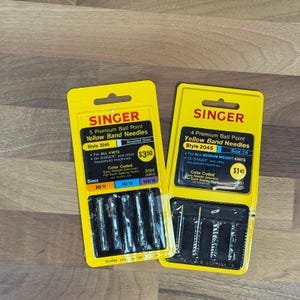 May include: Two yellow Singer packaging cards containing sewing machine needles. The left card holds five assorted premium ball point yellow band needles, style 2045, for all knits. The right card holds four size 14 premium ball point needles.