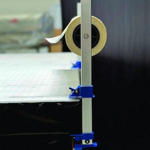 May include: A white roll of paper is mounted on a silver metal stand with blue clamps. The stand is attached to a black surface.