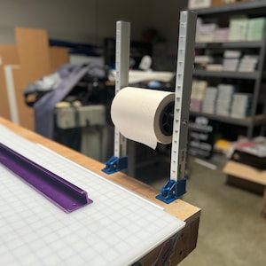 May include: A white roll of paper is mounted on a metal stand with blue and white accents. The stand is attached to a wooden table with a purple ruler and a white gridded surface.