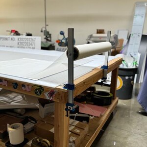 May include: A wooden work table with a white roll of material attached to two blue clamps. The roll is attached to a metal bar that is also clamped to the table. There are several other items on the table, including a roll of tape, a box of tape, and a bottle of water.