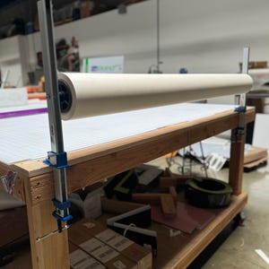 May include: A white roll of material is mounted on a wooden table with two blue and silver clamps. The table has a lower shelf and wheels. The roll is mounted on a metal bar with a handle.