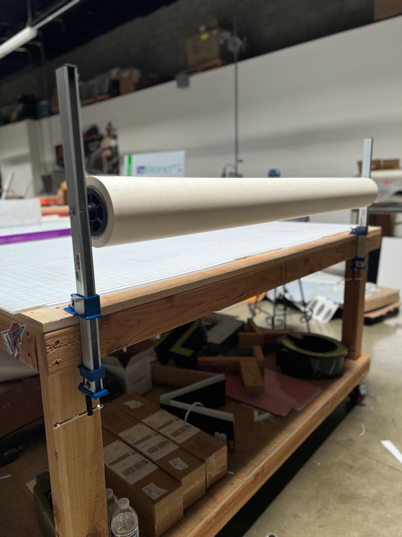 May include: A white roll of material is mounted on a wooden table with two blue and silver clamps. The table has a lower shelf and wheels. The roll is mounted on a metal bar with a handle.