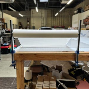 May include: A white roll of paper is mounted on a wooden table with two blue clamps. The table is covered with a white sheet of paper. There are various items on the table, including boxes, rolls of tape, and a clear water bottle.