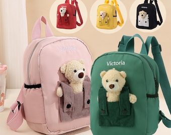 May include: Three pink, green, and yellow children's backpacks with teddy bear appliques. Each backpack has the word "Victoria" embroidered on the front.