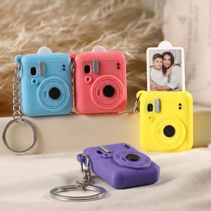 May include: A collection of colorful miniature camera keychains in blue, pink, yellow, and purple. Each keychain features a circular lens, a flash, and a silver chain with a keyring. One yellow camera holds a small photo.
