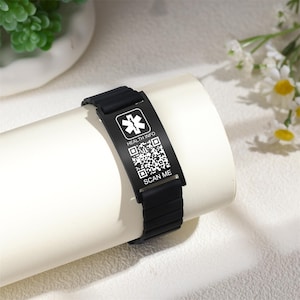 Może przedstawiać: Czarna medyczna bransoletka identyfikacyjna z prostokątną płytką z symbolem medycznym, "HEALTH INFO", kodem QR i tekstem "SCAN ME". Bransoletka jest prezentowana na białej powierzchni.