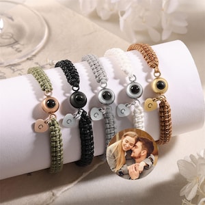 May include: Five friendship bracelets with a hidden photo compartment. Each bracelet is made with different colored string and has a small heart charm with the letter 'C' on it. The bracelets also have a small round charm with a black eye in the center.
