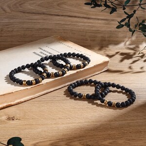 May include: Four black lava stone bracelets with gold beads and engraved lettering. The bracelets are stacked on top of a book.