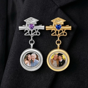 Puede incluir: Dos broches con temática de graduación. Uno es plateado con una gema púrpura, el otro dorado con una gema azul. Ambos tienen un gorro de graduación, el año "2026" y un marco de fotos circular.