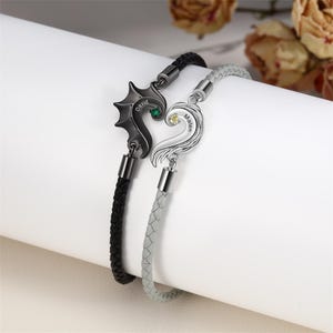 Op de afbeelding: Twee bijpassende armbanden, één zwart en één grijs, elk met een hartvormige hanger. De zwarte hanger heeft een drakenvleugelontwerp en de naam "David". De grijze hanger heeft de naam "Maren". Beide hebben gevlochten leren bandjes.