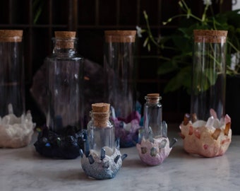 Puede incluir: Colección de botellas de vidrio con tapones de corcho, algunas con bases de cristal. Las botellas son de vidrio transparente con corchos naturales. Las bases de cristal son en tonos de blanco, azul, morado y rosa. Medidas en cm.