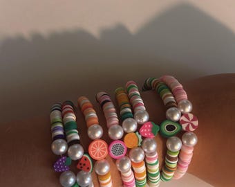 May include: A stack of colorful beaded bracelets with fruit charms. The bracelets are made with white, pink, green, orange, and yellow beads. The charms include a strawberry, watermelon, orange slice, avocado, and a purple fruit.